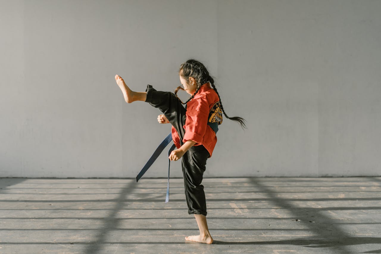 A young martial artist in a red uniform practicing a high kick indoors with natural lighting.